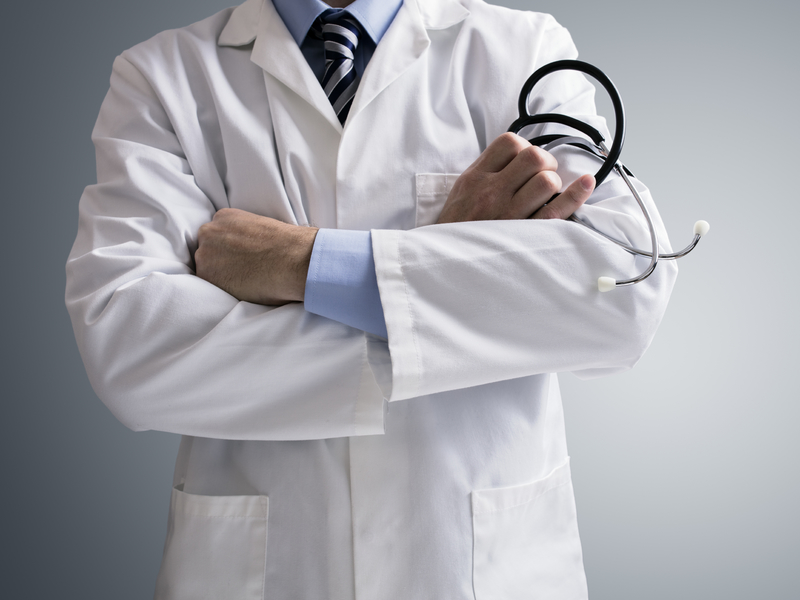 Homme les bras croisés portant une blouse de médecin, un stéthoscope dans la main.