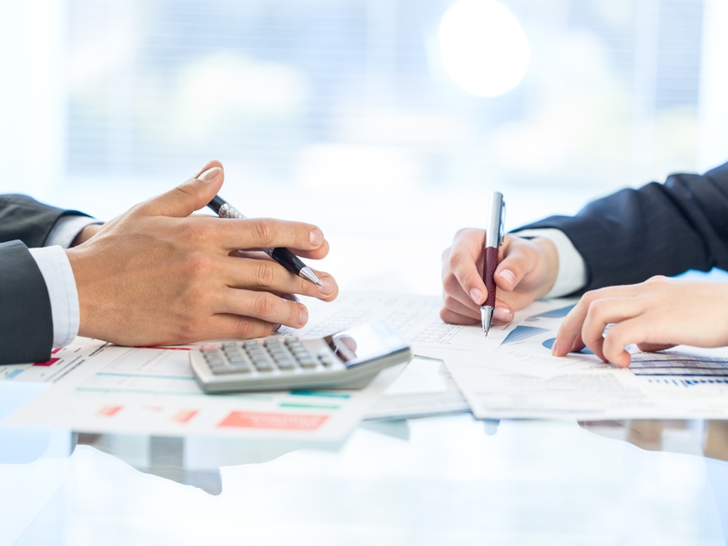 Deux hommes d'affaires écrivant sur des feuilles où on trouve des graphiques financier. Une calculette est posée devant l'un d'eux.