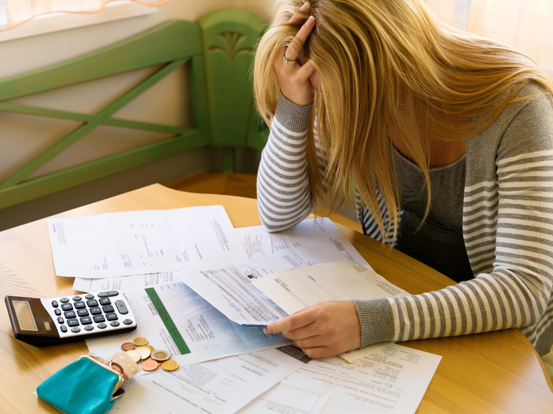 Une femme désespérée qui regarde une feuille. À côté d'elle on voit un portefeuille peu rempli et une calculette.