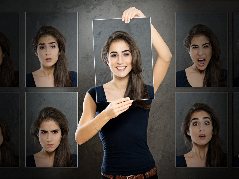 une femme qui tient une photo devant son visage représentant un visage souriant. On voit d'autres photos autour d'elle qui représentent d'autres émotions.