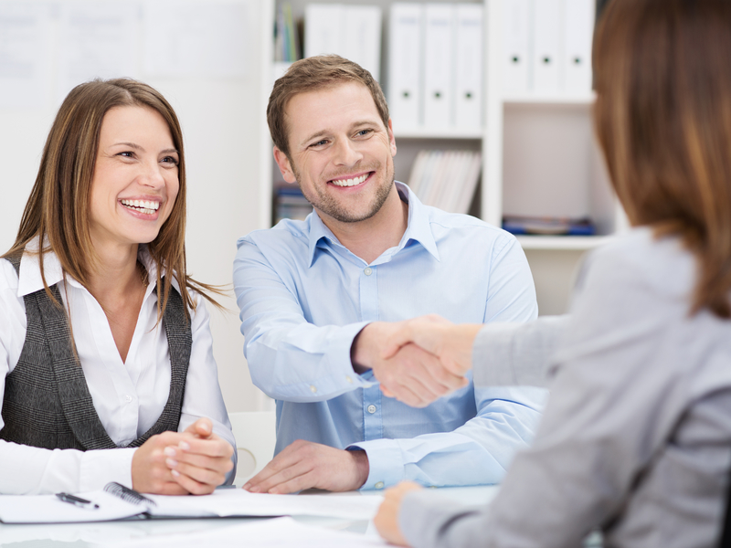 Un couple souriant devant un bureau. L'homme serre la main à une femme en tailleur qui est assise de l'autre côté de la table.