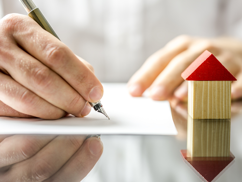 Un homme d'affaire signant un document sur une table, devant lui on voit une petite maison en bois.