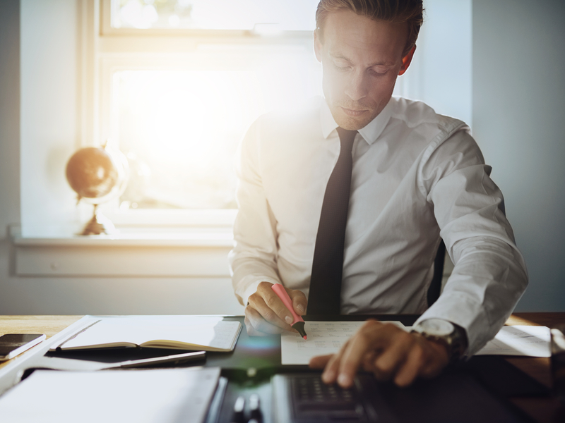 un comptable assis à un bureau. Il écrit sur des feuilles posés devant lui, son autre main pianote sur une calculatrice.