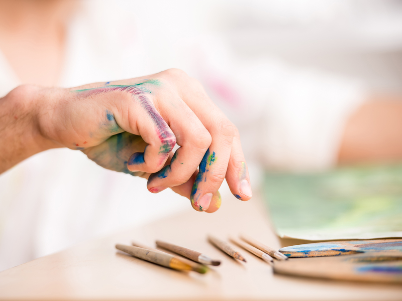 Une main pleine de peinture qui prend une aquarelle posée sur la table devant une feuille.