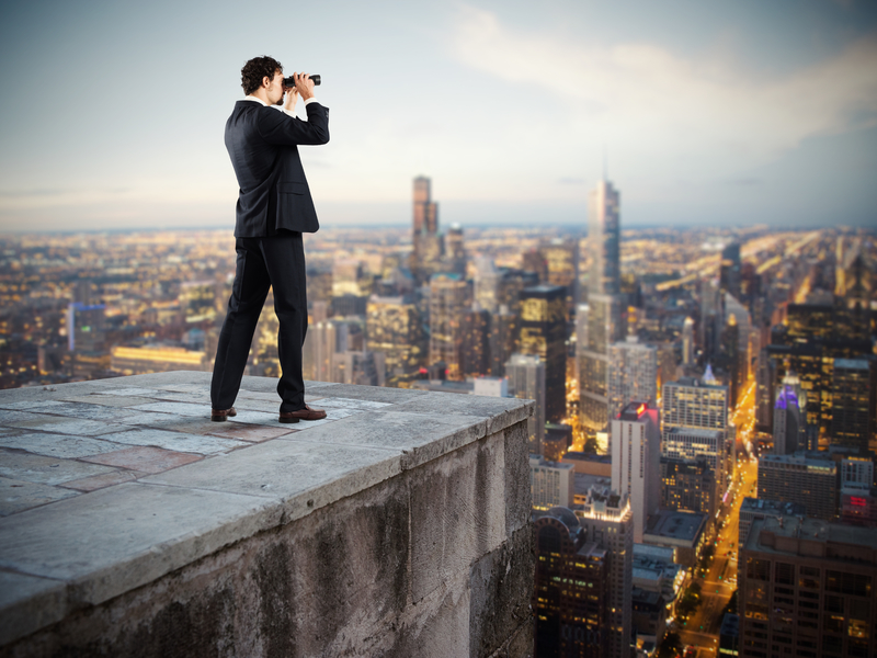 Un homme d'affaire avec des jumelles debout sur un toit d'immeuble observant l'horizon avec des jumelles.