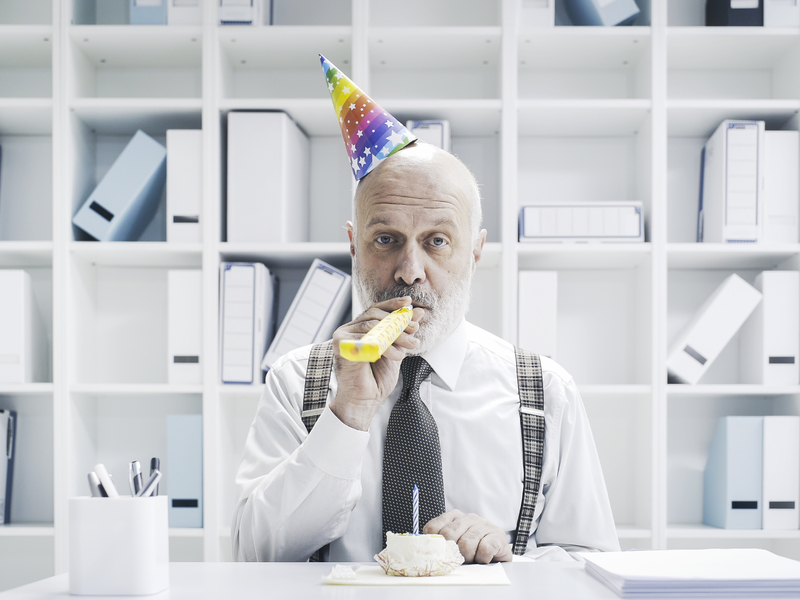 Un homme d'affaire avec un petit chapeau d'anniversaire et une trompette de fête, assis à un bureau devant un gâteau.