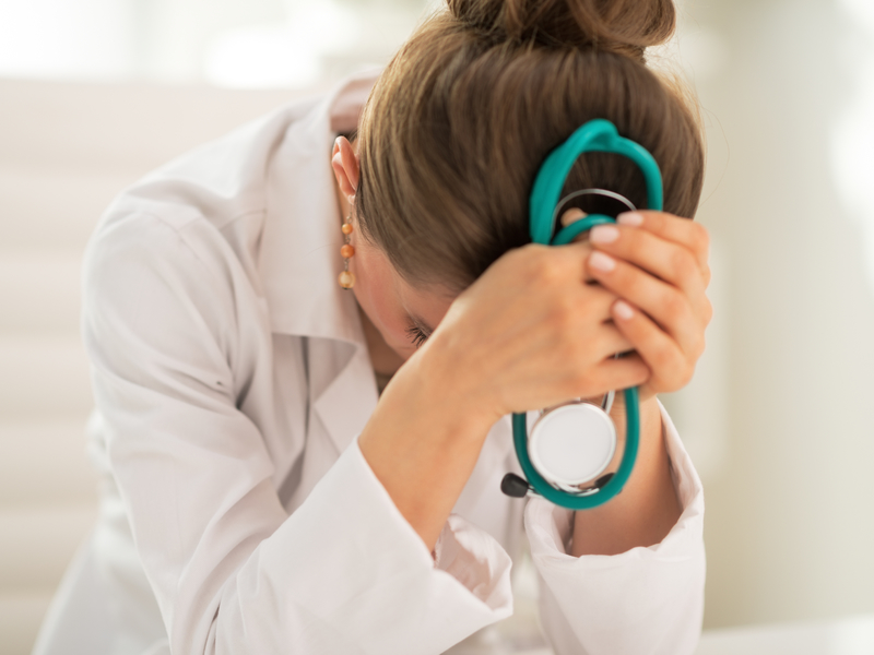 Une femme médecin qui se cache la tête dans les bras. Ses mains tiennent un stéthoscope. Elle a l'air stressée.