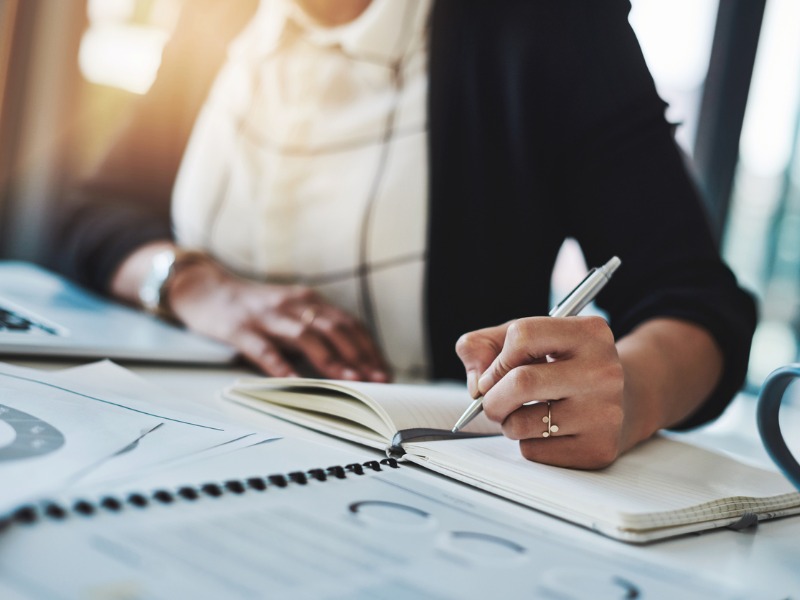 Une femme écrivant sur un cahier devant des graphiques financiers.