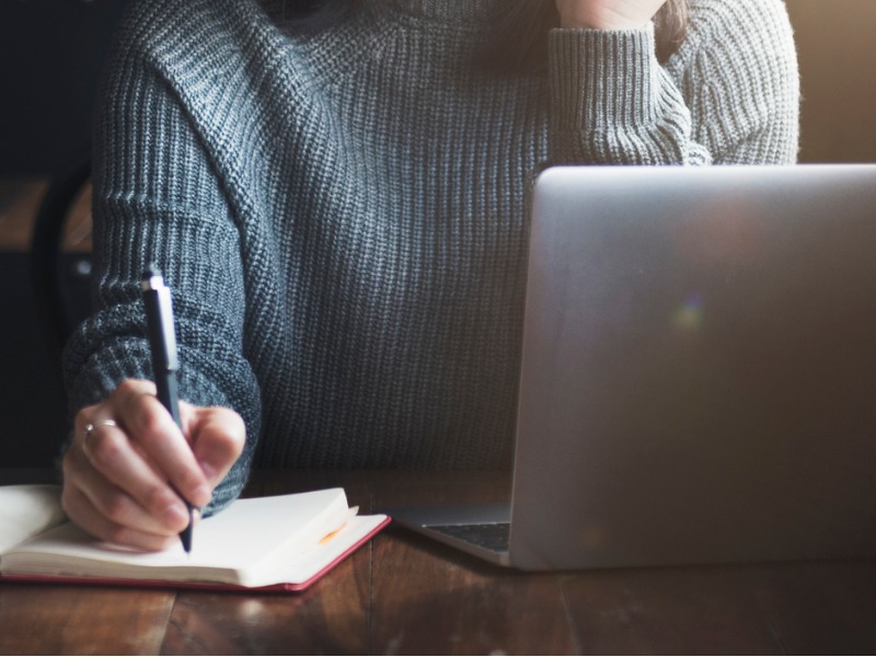 Une femme assise à un bureau devant un ordinateur. Elle écrit sur un bloc note.