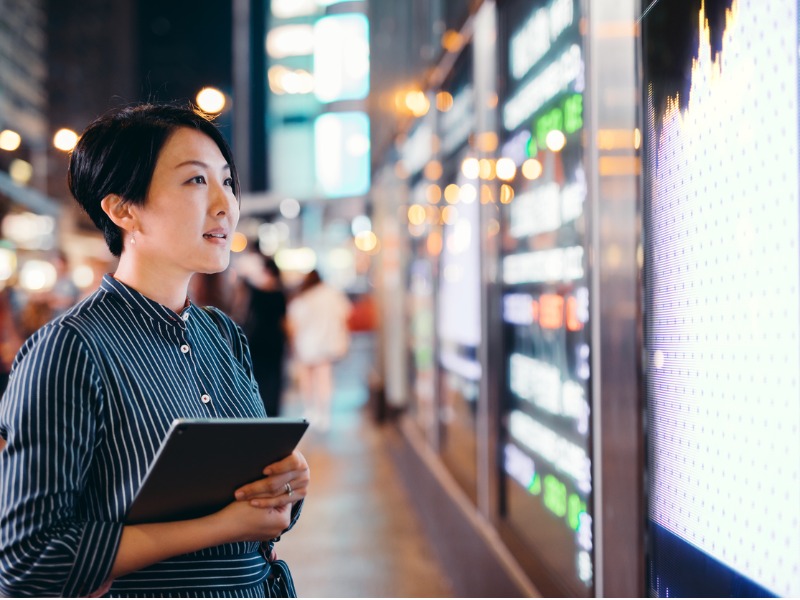 Une femme asiatique regardant un panneau représentant les cours du marché dans la rue.