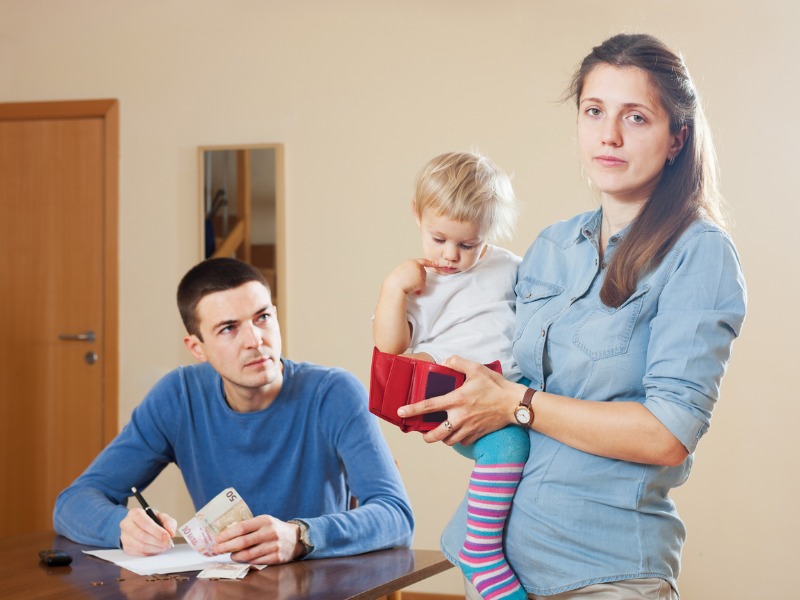 Une femme portant un bébé dans un bras, son portefeuille dans la main à côté de son mari qui écrit sur des papiers sur une table.