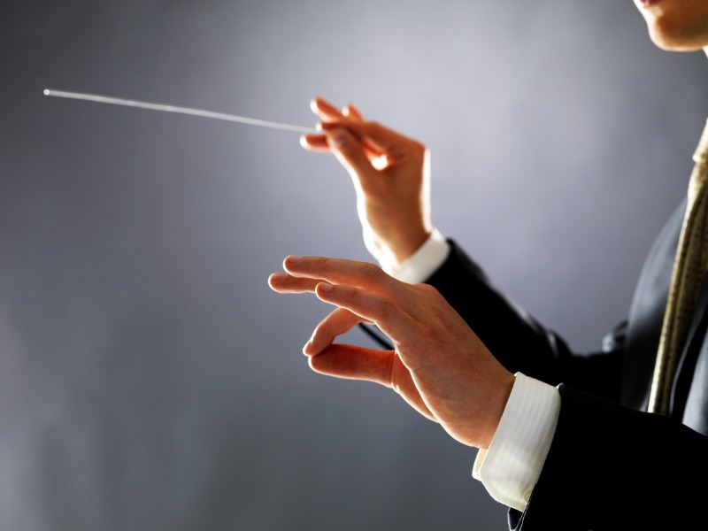Une photo d'un homme en costume, dont on voit les deux mains prête à diriger un orchestre. Il tient une baguette dans la main droite.