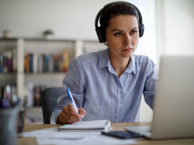 Une femme assise à un bureau devant un ordinateur. Elle porte des écouteurs et semble prendre des notes.