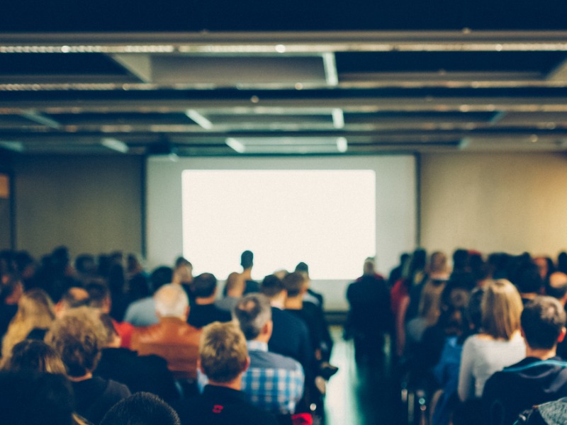 Une salle de conférence remplie par le public qui attend assis devant un écran blanc.