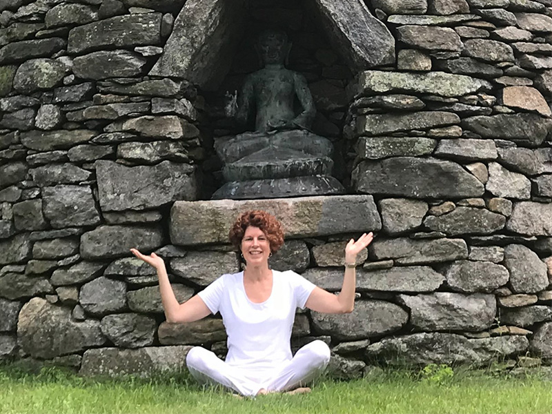 Photo de Mary Hagerman assise en tailleur devant un mur de pierre dans lequel on voit un buddha en tailleur.