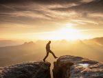 Homme en tenue de randonnée marchant entre deux gros rochers sur fond de coucher de soleil.