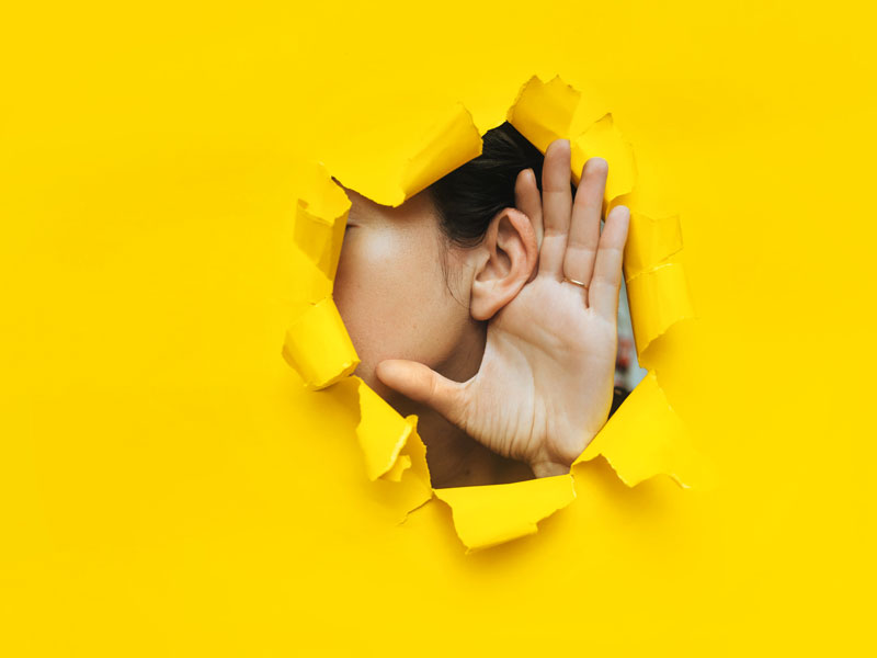 Gros plan de l'oreille et de la main d'une femme à travers un trou déchiré dans le papier. Fond jaune, espace réservé à la copie. Concept d'écoute, d'espionnage, de ragots et de tabloïds.