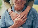 Une jeune femme placée derrière une personne âgée avec les bras posés sur ses épaules, dans un geste très tendre. L'homme âgé tient une des mains de la jeune femme.