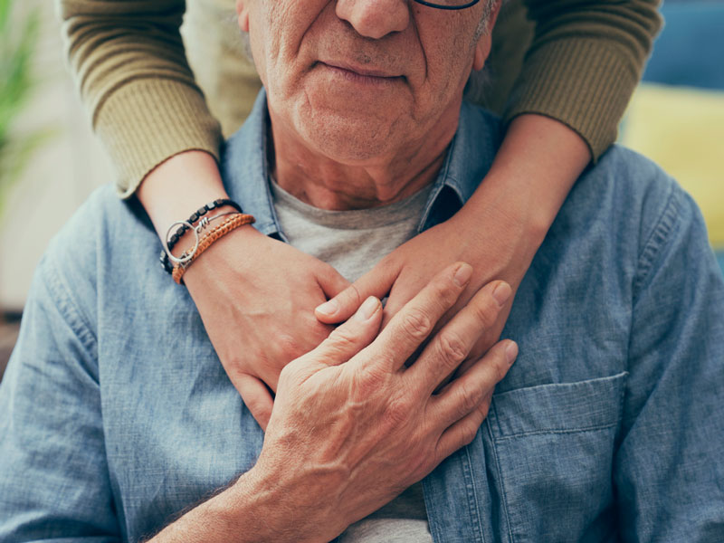 Une jeune femme placée derrière une personne âgée avec les bras posés sur ses épaules, dans un geste très tendre. L'homme âgé tient une des mains de la jeune femme.