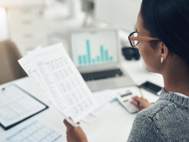 Femme assise à un bureau avec des feuilles et des graphiques, travaillant sur ordinateur portable et calculatrice.