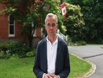 Portrait de Mark Carney à l’extérieur, vêtu d’une veste sombre et d’une chemise blanche. En arrière-plan, un drapeau canadien flotte devant un bâtiment en brique rouge entouré de verdure. L’image reflète un message institutionnel ou patriotique.