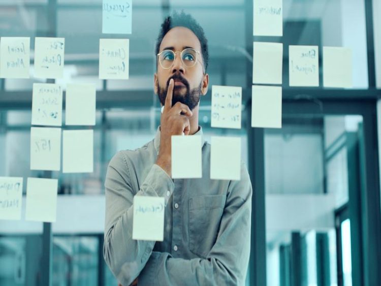 Un homme barbu avec lunettes réfléchit devant une vitre remplie de post-its, en pleine session de réflexion stratégique.