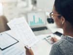 Femme concentrée sur des documents financiers devant un écran d’ordinateur.