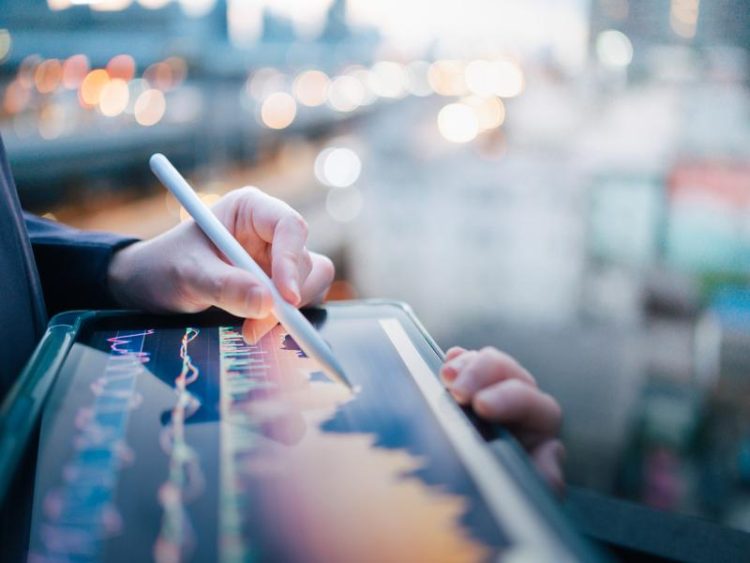 Main tenant un stylet au-dessus d’une tablette affichant un graphique boursier interactif, en contexte urbain flou.