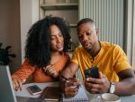 Jeune couple assis à une table, examinant leurs finances avec un téléphone.