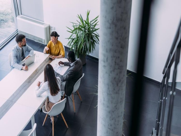 Vue d’en haut d’une réunion d’affaires dans une salle moderne. Quatre personnes discutent autour d’une table, documents et ordinateurs devant eux.