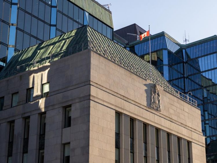 Façade du bâtiment moderne de la Banque du Canada, avec un toit vert, le drapeau canadien et des reflets sur les vitres.