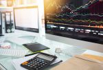 Écrans d’ordinateur affichant des graphiques financiers complexes avec un bureau de travail en premier plan (calculatrice, documents). Représentation d’un poste de travail d’analyste financier ou trader.