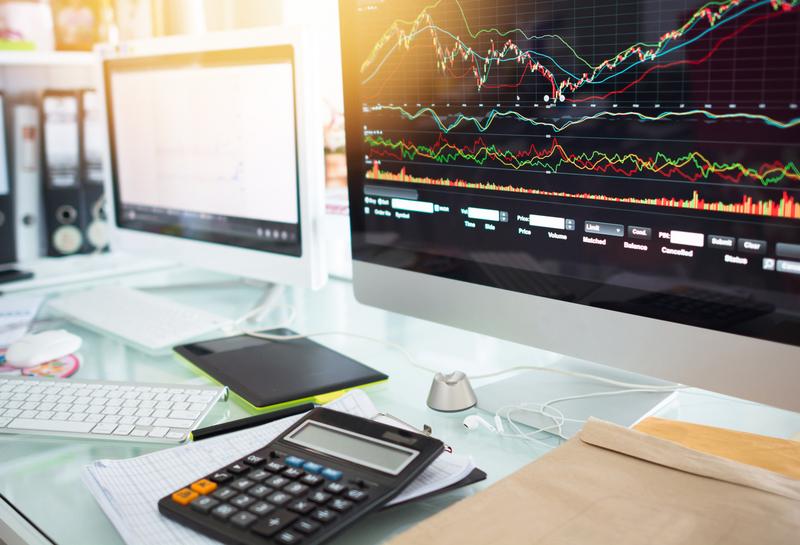 Écrans d’ordinateur affichant des graphiques financiers complexes avec un bureau de travail en premier plan (calculatrice, documents). Représentation d’un poste de travail d’analyste financier ou trader.