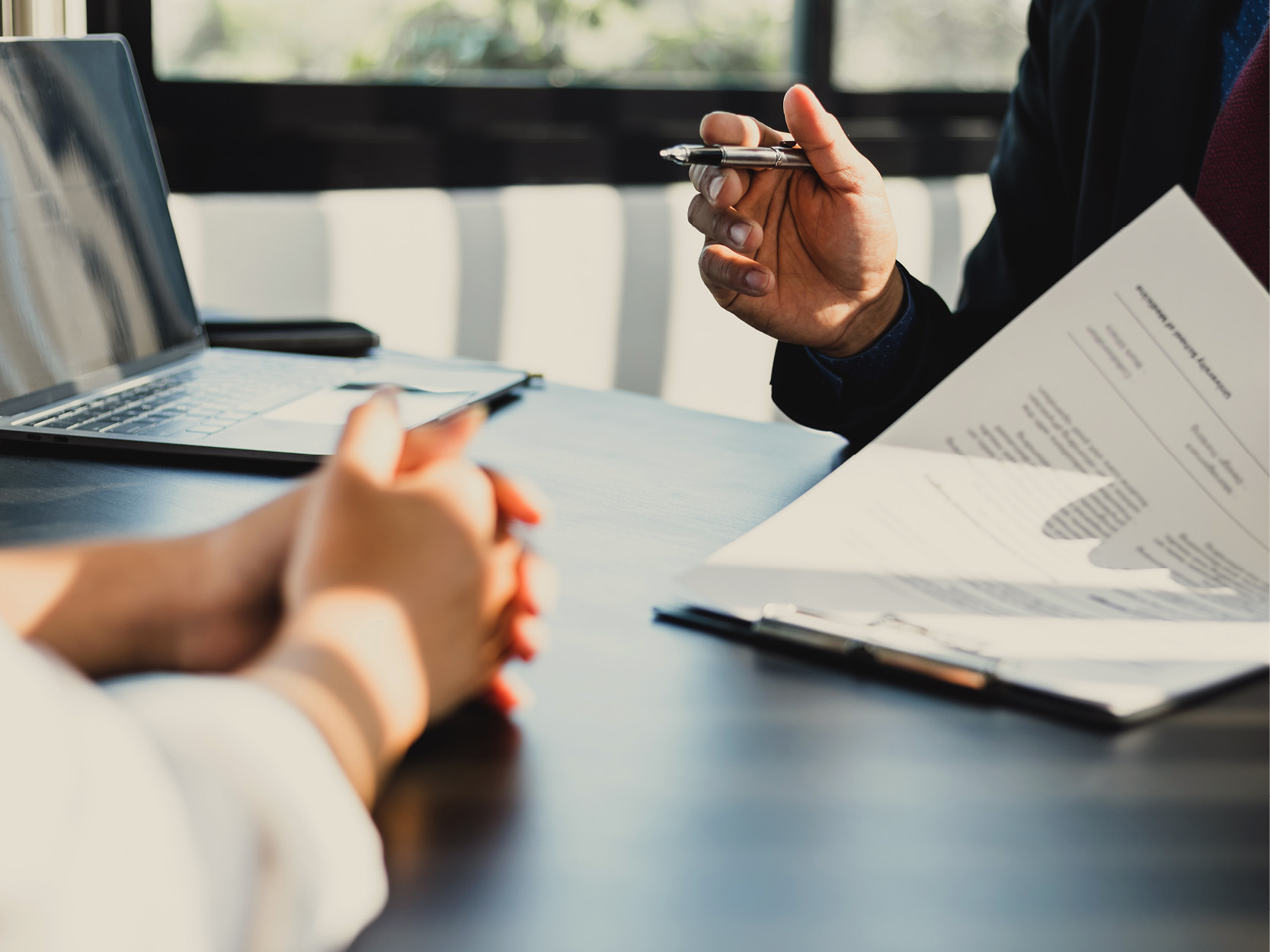 Rencontre entre un conseiller financier et un client autour d'une table de bureau moderne. Le conseiller tient un stylo et examine un rapport financier imprimé contenant des tableaux et des graphiques, tandis que le client écoute, les mains jointes. Un ordinateur portable ouvert est posé sur la table, la lumière du soleil pénétrant par de grandes fenêtres.