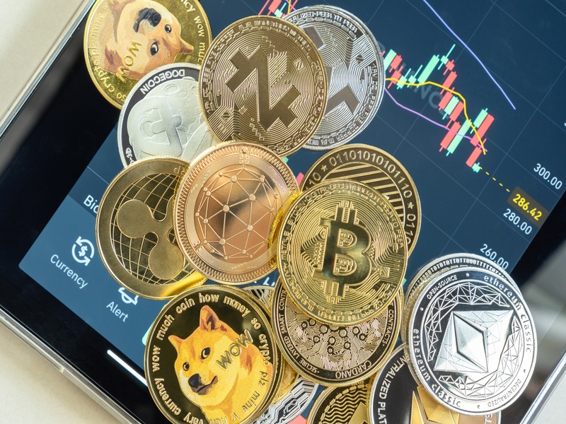 Collection de cryptomonnaies physiques, dont Bitcoin et Ethereum, posées sur un écran affichant des graphiques de trading. Illustration du marché des cryptos, de la blockchain et de l’investissement numérique.