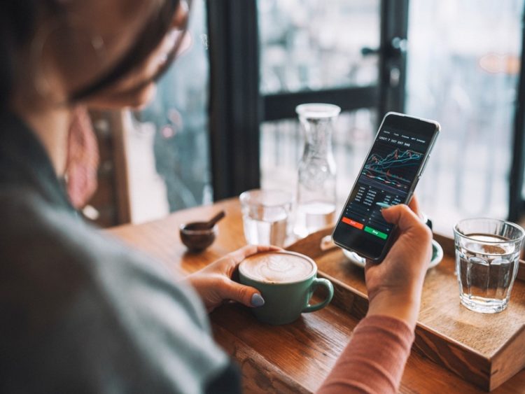 Personne consultant une application de trading sur smartphone tout en tenant une tasse de café, illustrant l’investissement mobile, le trading en ligne et la gestion financière moderne au quotidien.