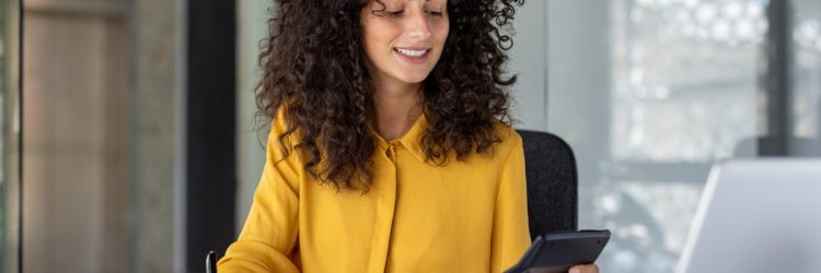 Jeune professionnelle souriante travaillant à son bureau, consultant des documents financiers avec une calculatrice et un ordinateur portable.