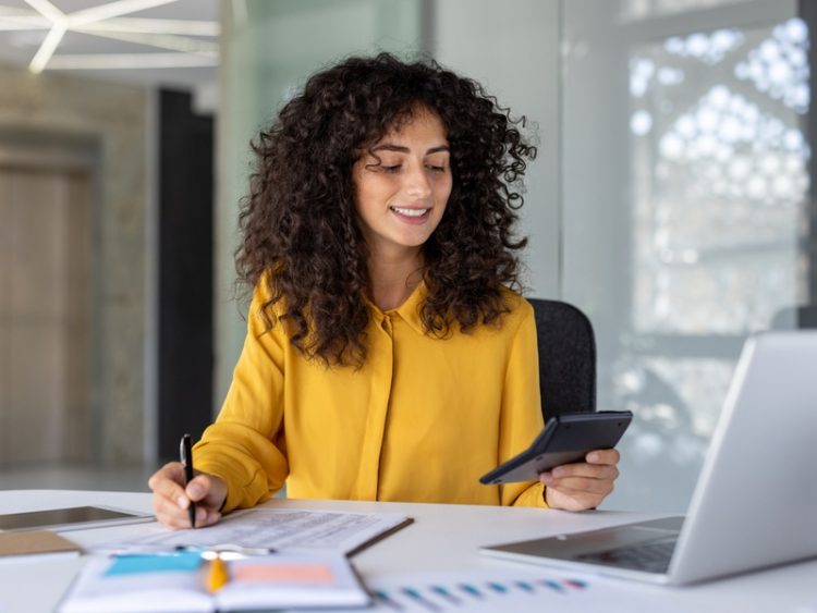 Jeune professionnelle souriante travaillant à son bureau, consultant des documents financiers avec une calculatrice et un ordinateur portable.