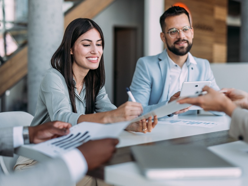 Une équipe de professionnels est réunie autour d’une table dans un environnement de bureau moderne et lumineux. Une femme souriante signe ou consulte un document pendant qu’un collègue tient une tablette numérique. D’autres participants échangent des documents, illustrant une réunion collaborative, un entretien ou une discussion d’affaires dans une ambiance conviviale et professionnelle.